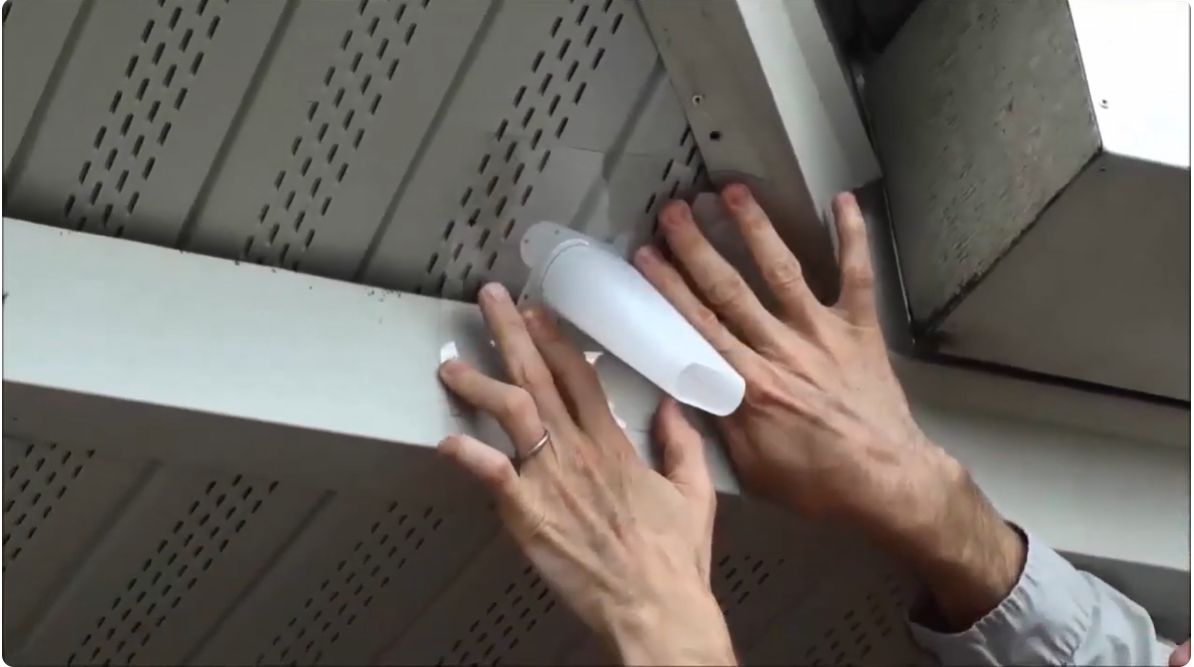 Two hands holding up a long plastic cone with tapered ends. The cone is positioned in the area under the eaves of a house.